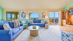 Un ampio soggiorno con una grande finestra che offre vista sulla spiaggia, un divano blu, un tavolino in vetro e un mobile in legno con oggetti decorativi.