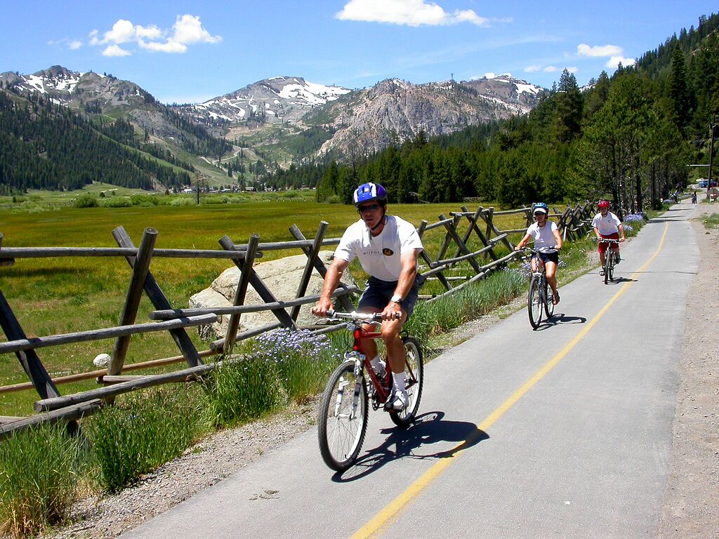 A paved path with cyclists, surrounded by a wooden fence and a mountainous landscape.