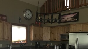 Ett kök med en köksö i mitten, två barstolar, träskåp, en klocka och en hängande lampa.