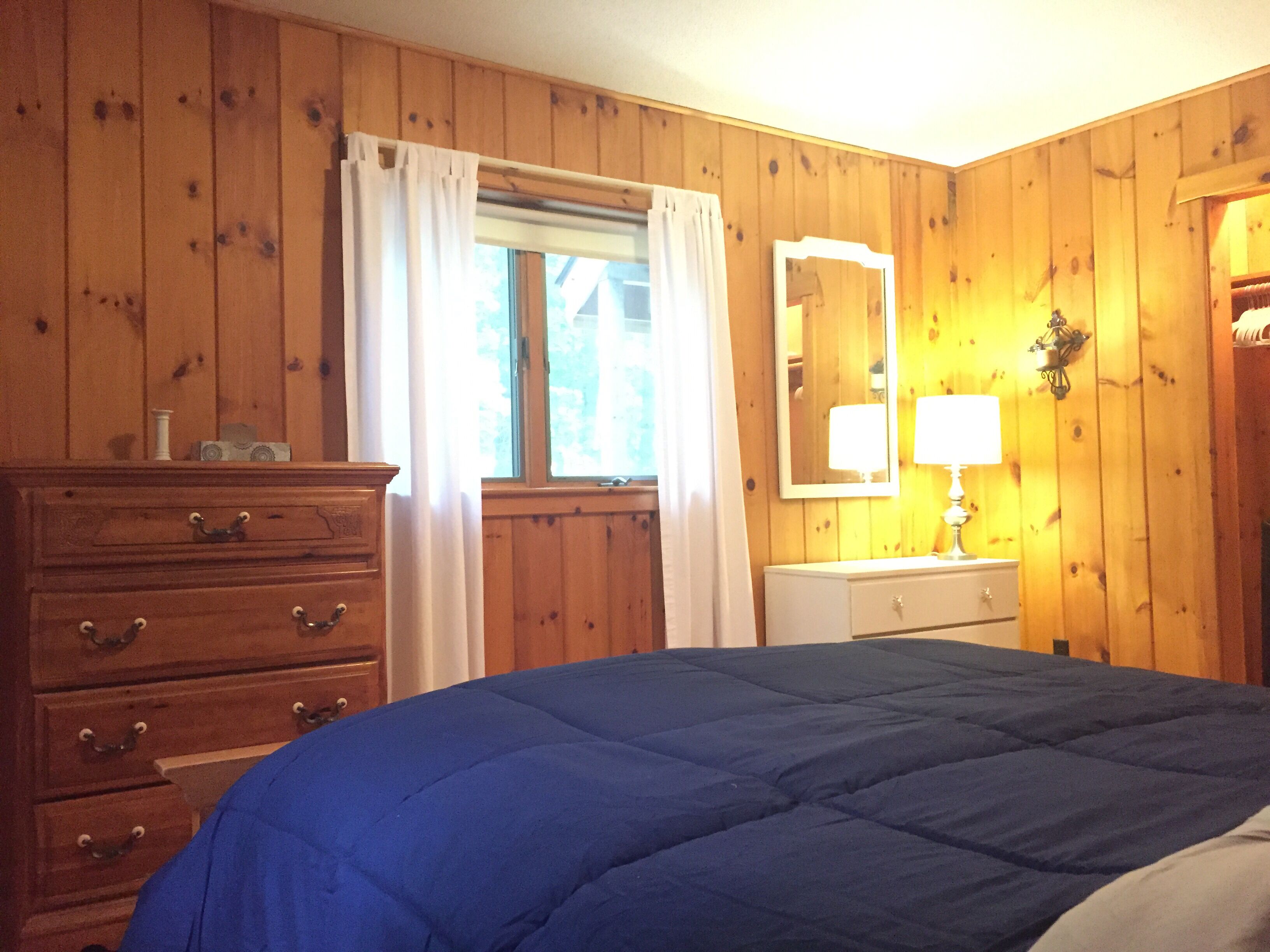 Una habitación con paneles de madera, una cama con cubrecama azul, un aparador con cajones, una ventana con cortinas, un espejo y una lámpara sobre una mesita de noche.