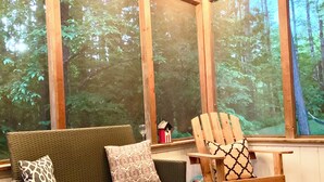 Une pièce chaleureuse avec un canapé en osier, une chaise en bois et une petite table. De grandes fenêtres offrent une vue sur la forêt.