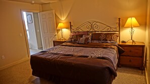 Quarto com uma cama grande, duas mesas de cabeceira com luminárias, um criado-mudo de madeira e uma porta que dá para outro cômodo.