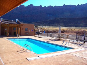 A swimming pool with a view of a mountain range, a covered patio area, and a fenced outdoor seating area.