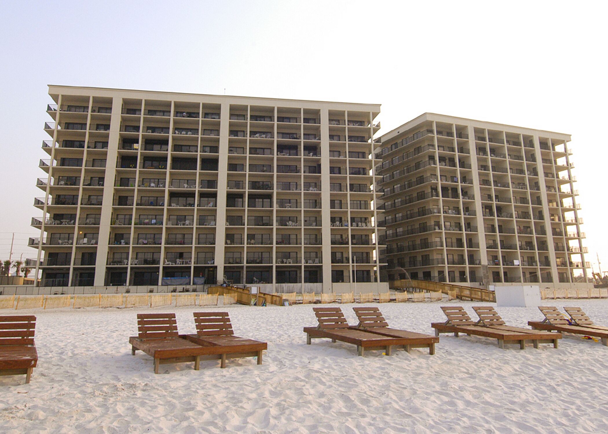 A multi-story hotel with balconies, located on a sandy beach with wooden lounge chairs.