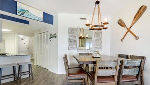 A dining area with a wooden table and chairs, a chandelier, and a wall-mounted paddleboard. - End of Year Special Unit #413 Rates-Beachfront Beautifully Renovated  2/2Sleeps8 (Orange Beach)