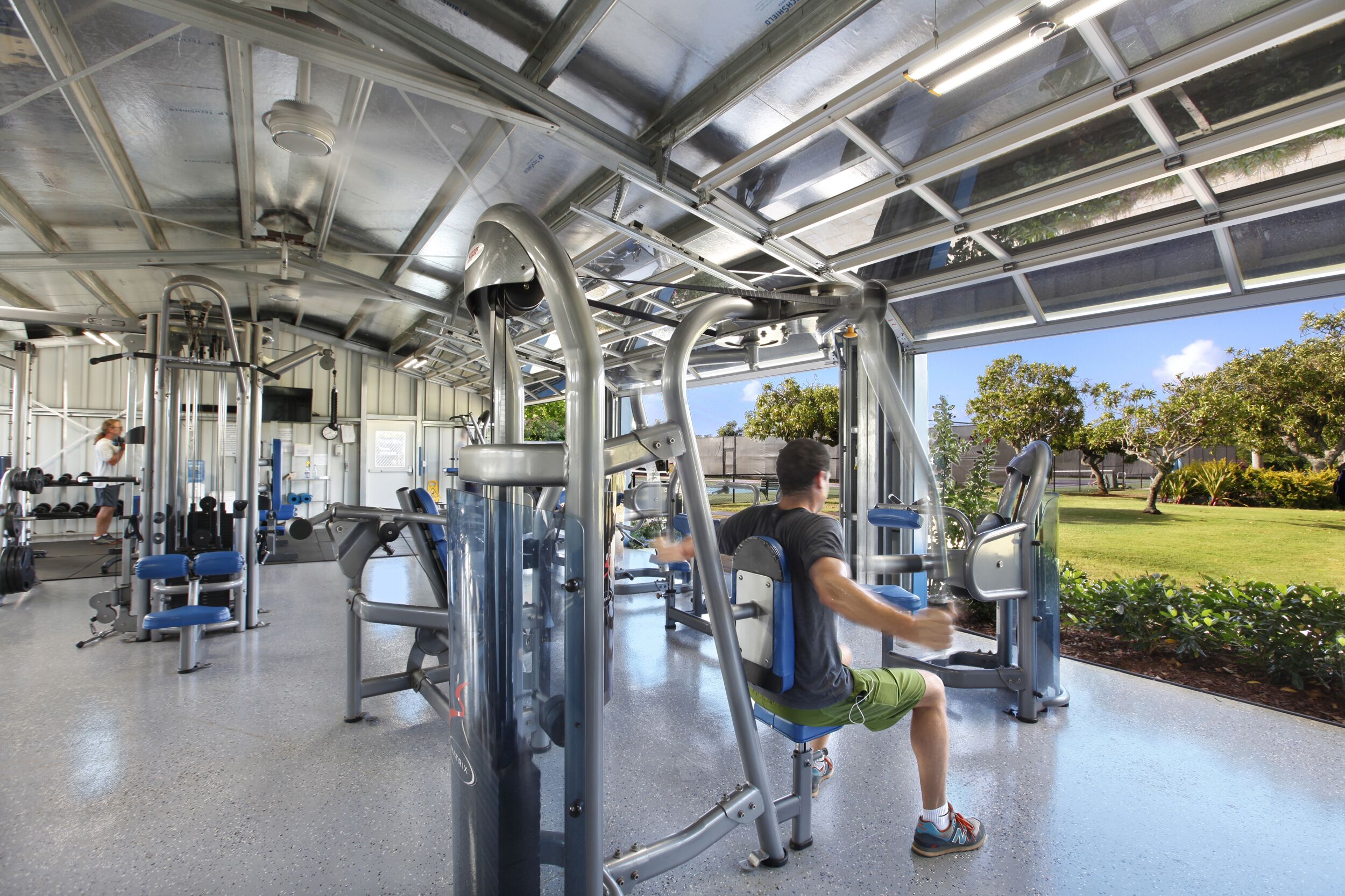 Ein Fitnessstudio mit verschiedenen Trainingsgeräten, einem überdachten Bereich und Blick auf eine grüne Landschaft.