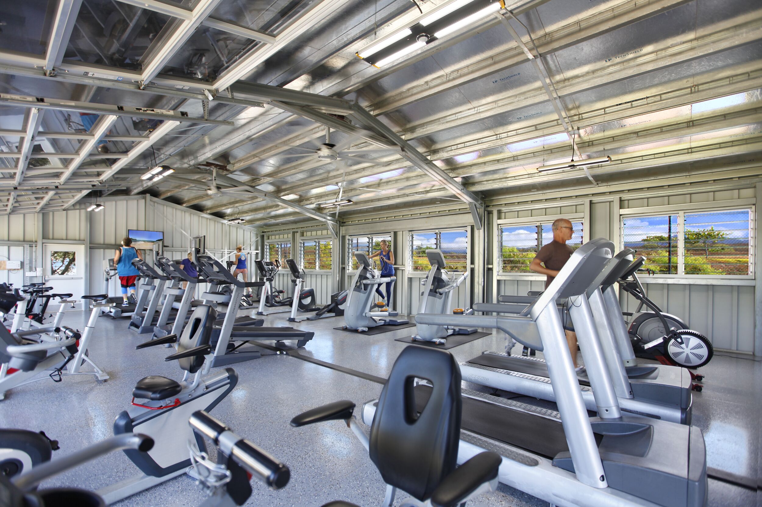Un gimnasio con diversas máquinas de ejercicio, ventanas amplias y vistas al exterior.