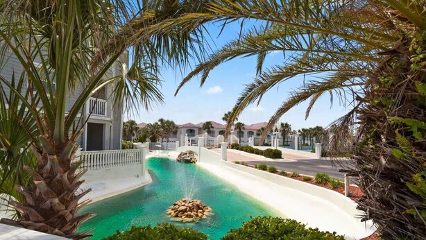 A pool with a rock feature surrounded by palm trees and a building with a white facade. - Villas of Ocean Gate #106 Ocean side 2 bedroom, pool view, pet friendly (St Augustine)