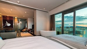 A modern hotel room with a large bed, a sitting area with a chair, a desk, and a large window offering a city view.