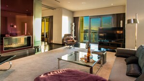 A modern living room with a glass coffee table, a sofa, a leather chair, a fireplace, and a large TV.
