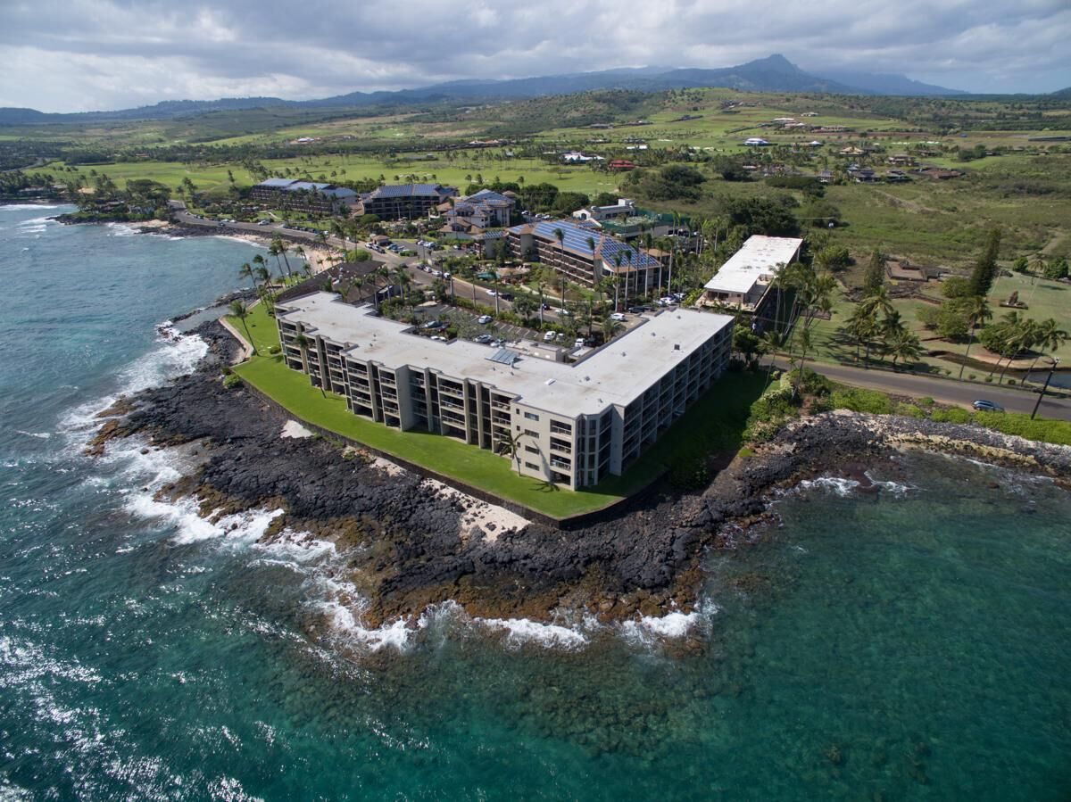 A coastal hotel with a large building, a parking area, and a view of the ocean and hills.
