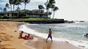 Ein Strand mit Palmen, ein Haus im Hintergrund und Menschen im Sand und im Wasser.