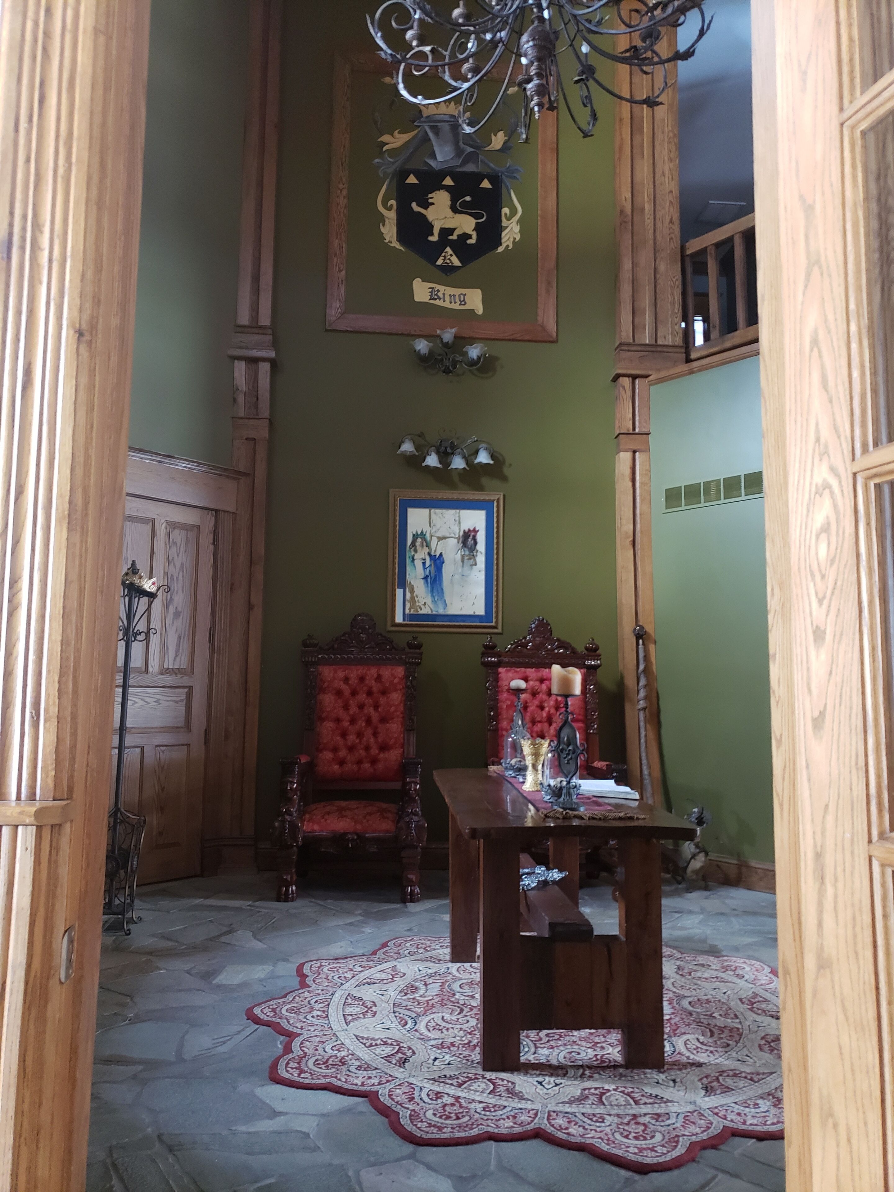 A room with a wooden floor, a red upholstered chair, a wooden table, a chandelier, and a coat of arms on the wall.