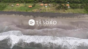 An aerial view of Playa Tesoro in Nicaragua, featuring a beachfront with waves, a grassy area with several small buildings, and dense greenery in the background. - Your Nicaragua Beachfront Hideaway – 2BR at Playa Tesoro (Leon)