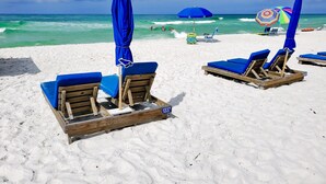 A beach with wooden lounge chairs and blue umbrellas, a clear sky, and the ocean in the background. - Luxury Ocean Front End Unit. Unobstructed sunset views! Beach service included! (Panama City Beach)