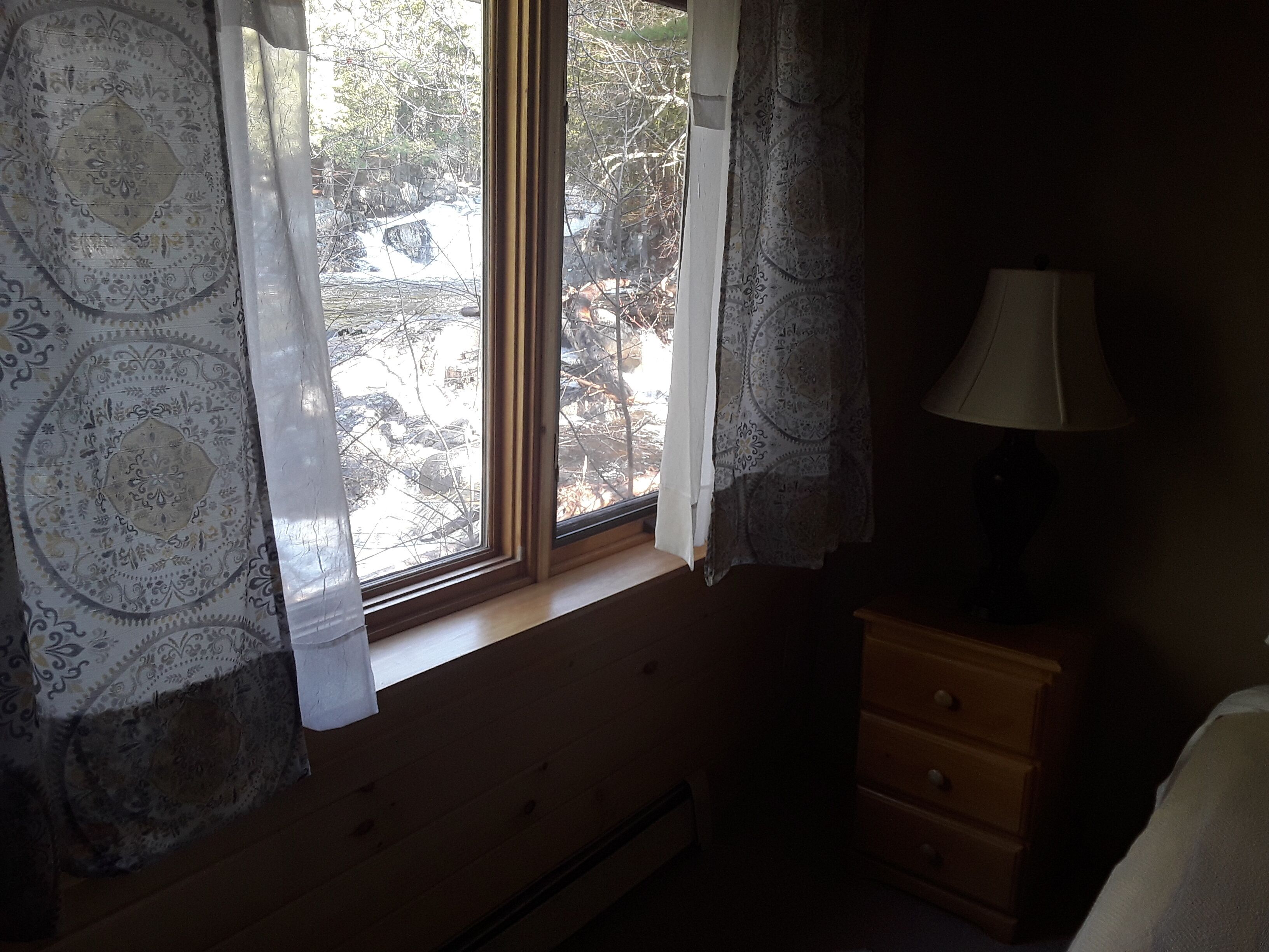 A room with a window, curtains, a lamp, and a wooden dresser.
