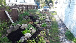 Un jardin agrémenté de diverses plantes et de rochers, une clôture en bois et un véhicule garé au fond.