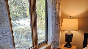 Une chambre à coucher avec un lit en bois, une table de chevet avec une lampe, une fenêtre avec des rideaux et une vue sur les arbres et les rochers à l’extérieur.