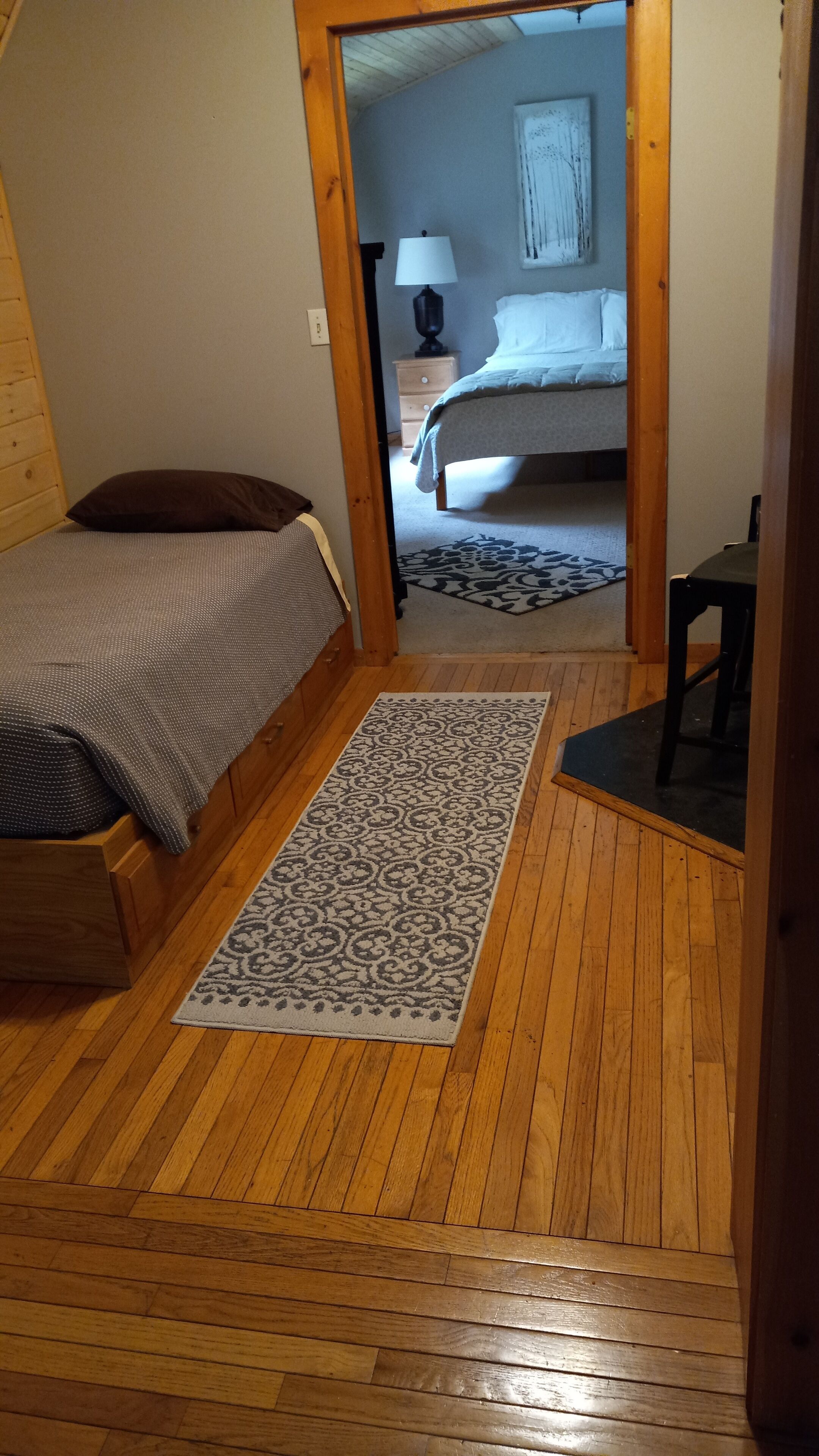 A bedroom with a wooden floor, a bed with a patterned bedspread, a nightstand with a lamp, and a painting on the wall.