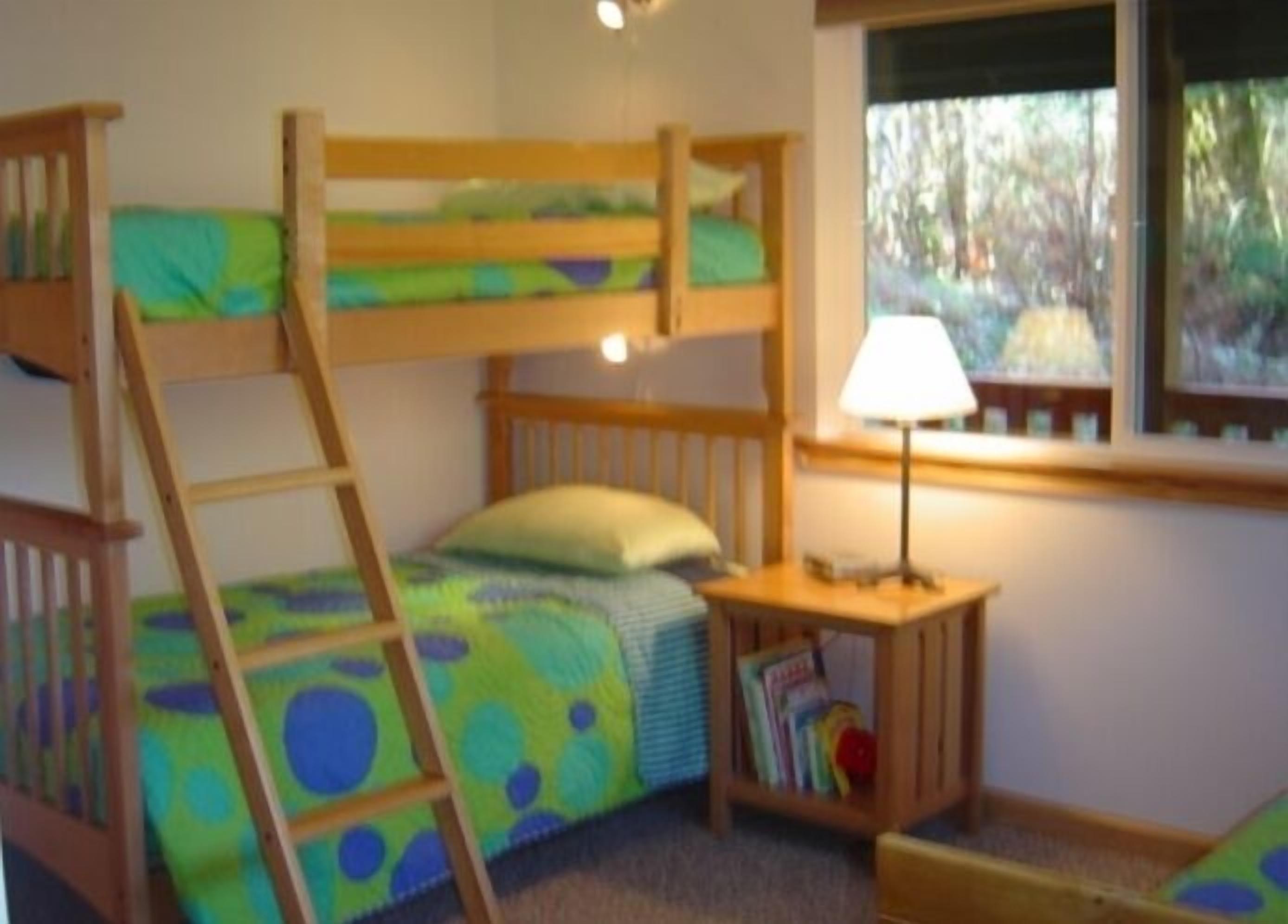 Un lit superposé avec une échelle, une table de chevet avec une lampe et une fenêtre donnant sur les arbres.