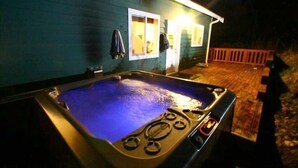 Un jacuzzi installé à l’extérieur d’une maison, avec une terrasse en bois et un petit porche.