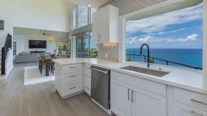 A modern kitchen with white cabinetry, a built-in dishwasher, and a large window overlooking the ocean. - Honu Awa: Upscale Oceanfront Home w/ Pool, AC & Cliffside Views (Princeville)