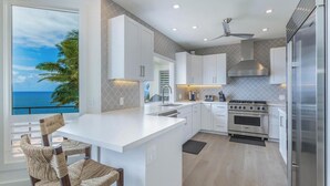 A modern kitchen with white cabinetry, a large island, stainless steel appliances, and a view of the ocean. - Honu Awa: Upscale Oceanfront Home w/ Pool, AC & Cliffside Views (Princeville)