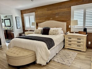 A bedroom with a wooden wall, a bed with a headboard, bedside tables, a lamp, a nightstand, and a mirror.