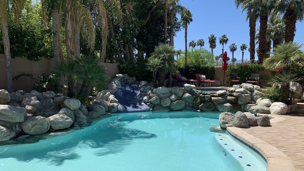 A clear blue pool with a waterfall feature, surrounded by large rocks and palm trees. - Amazing large Pool & Beautiful Desert Oasis in the Palm Desert area. (Palm Desert)