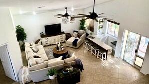 A spacious living area with a sofa, coffee table, and a dining area with stools. There are ceiling fans, a TV, and a window with a view. - Amazing large Pool & Beautiful Desert Oasis in the Palm Desert area. (Palm Desert)