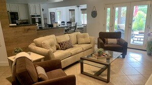 A living room with a beige sofa, a brown armchair, and a glass coffee table. There is a kitchen in the background with white cabinets and a refrigerator. - Amazing large Pool & Beautiful Desert Oasis in the Palm Desert area. (Palm Desert)