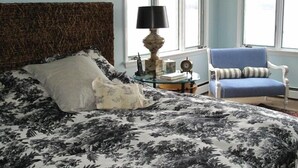 Een slaapkamer met een groot bed, een nachtkastje met een lamp, een klok en een blauwe stoel bij een raam.