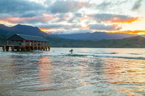 A pier extending into calm waters with a person surfing, surrounded by mountains and a sunset.