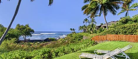 A lush garden with a wooden fence, two lounge chairs, and a view of the ocean.