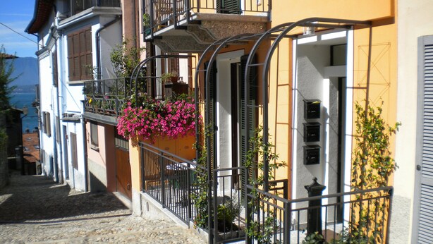 Une ruelle étroite pavée, bordée de bâtiments de chaque côté, avec des balcons ornés de jardinières et de plantes en pot.