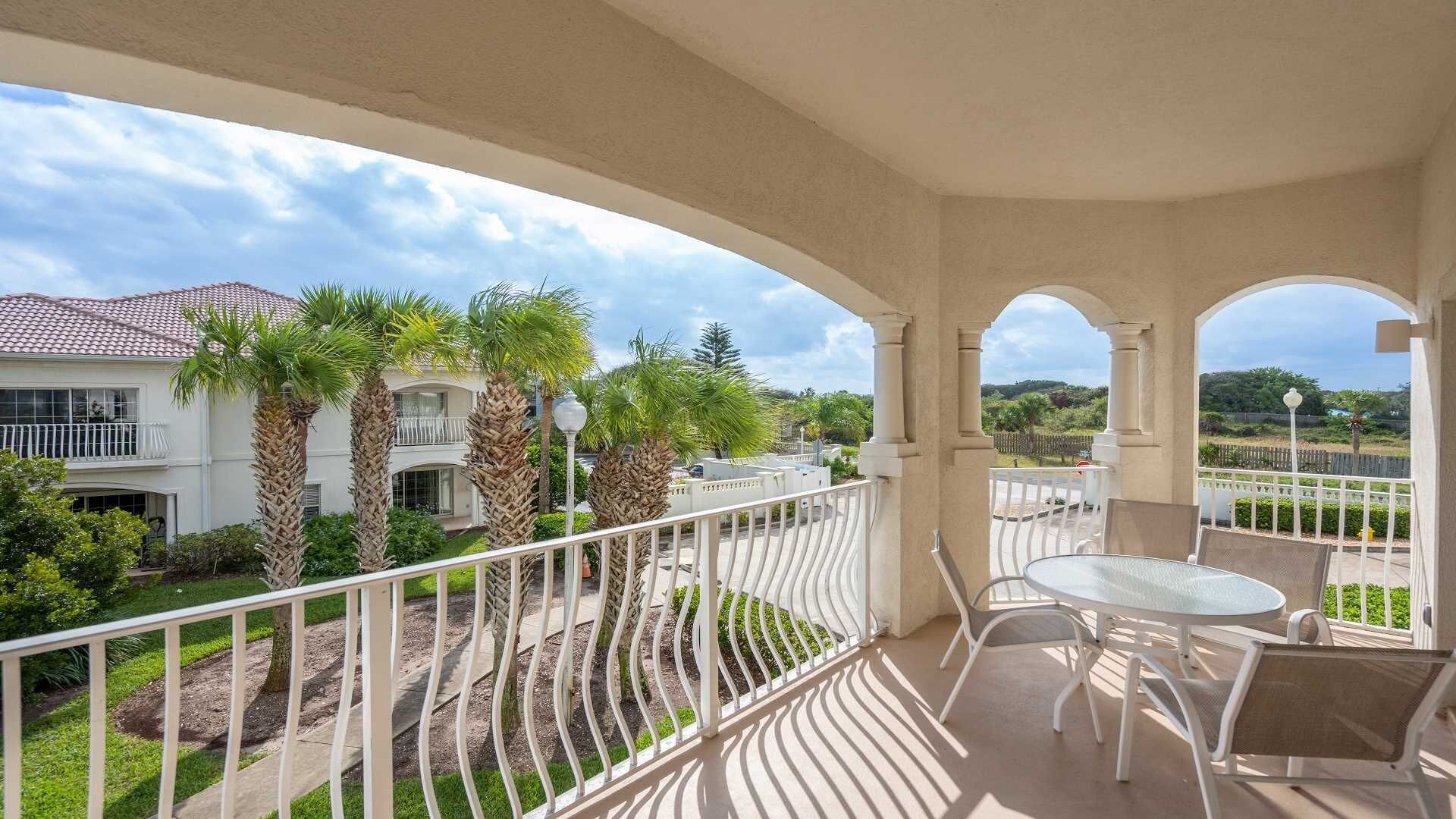 A balcony with a table and chairs, overlooking a residential area with palm trees and a house with a tiled roof.