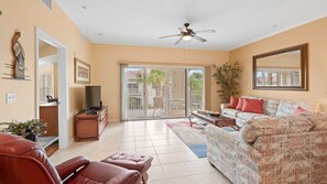 A spacious living room with a sofa, a coffee table, and a TV stand. There is a sliding glass door leading to an outdoor patio area.