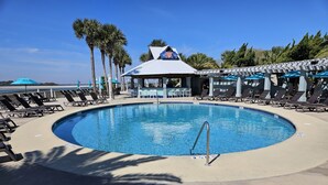 A pool with lounge chairs and umbrellas, surrounded by palm trees and a building with a blue roof. - Enjoy our new 16x25 screened in porch on Ocean Point Golf course (Fripp Island)
