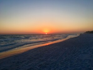 A beach with a sunset over the ocean, sandy shore, and a few people in the distance.