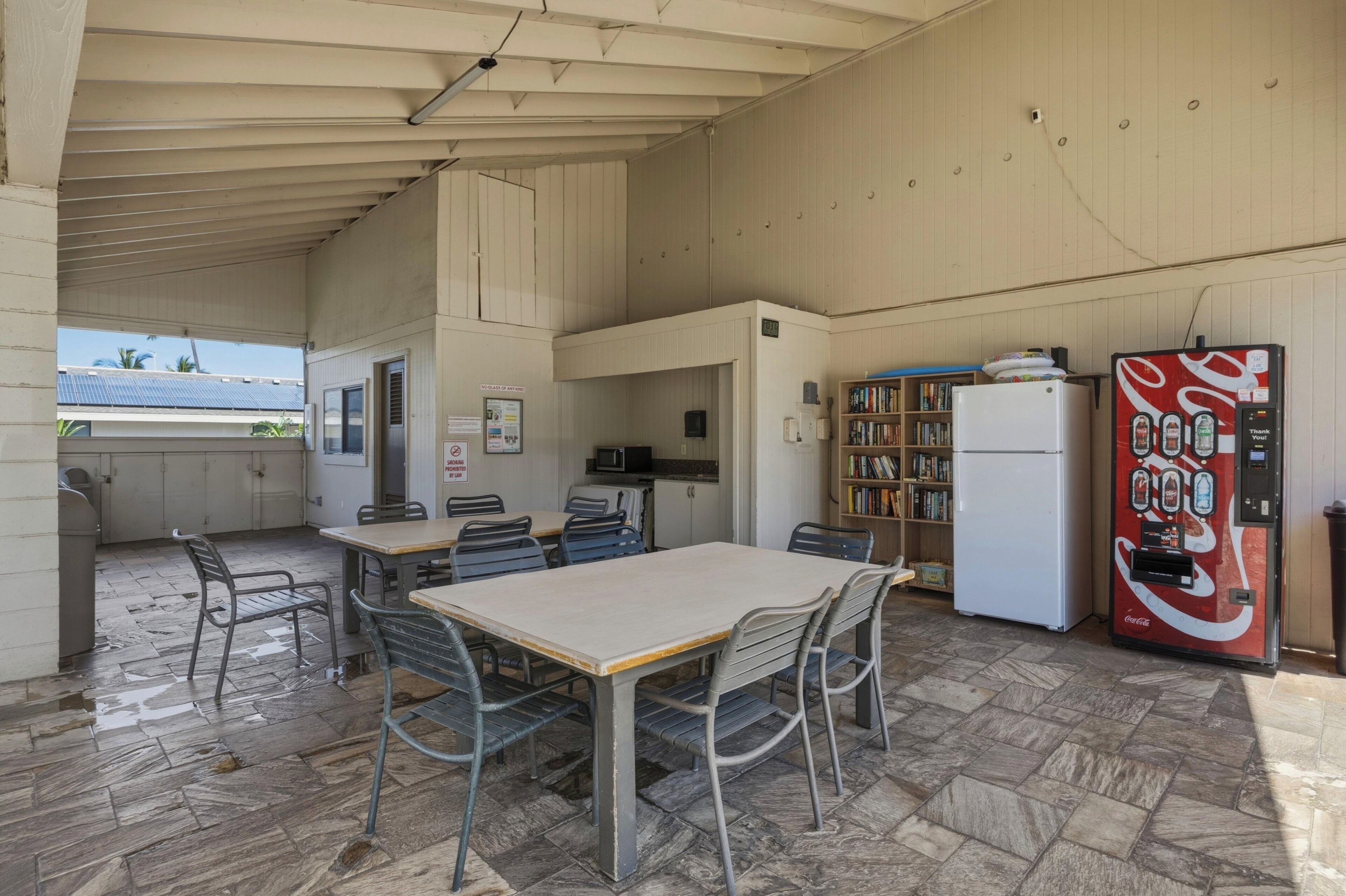 A covered outdoor area with a table and chairs, a refrigerator, a bookshelf, and a Coca-Cola vending machine.