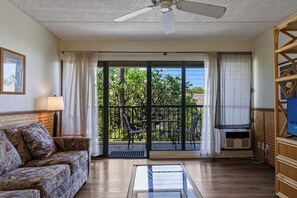 A room with a ceiling fan, a sofa, a TV, a balcony with chairs, and a view of greenery.