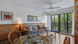 Sala de estar con sofá, sillas, una mesa de vidrio, ventilador de techo y ventanas amplias con vista a follaje.