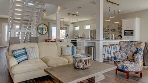A spacious living room with a sofa, armchair, and coffee table. A kitchen with white cabinets and a dining area with a table and chairs are visible in the background.