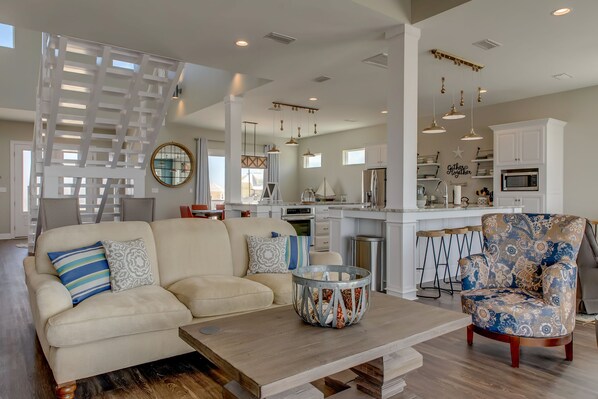 A spacious living room with a sofa, armchair, and coffee table. A kitchen with white cabinets and a dining area with a table and chairs are visible in the background.