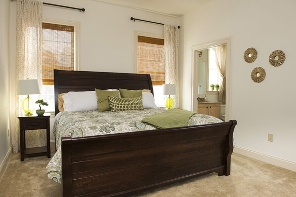 A bedroom with a large bed, two bedside tables, a nightstand, a mirror, and a window with blinds.