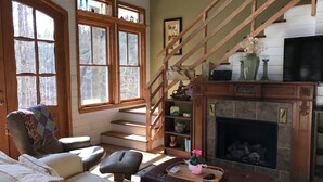 Uma sala de estar aconchegante com lareira, escada e vista para árvores através de grandes janelas.