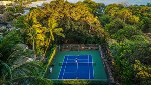Ein Tennisplatz im Freien, umgeben von üppiger Vegetation und einem malerischen Blick auf Strand und Berge.