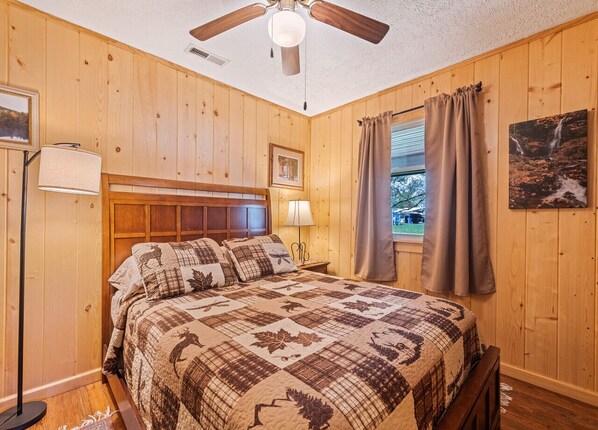 A wooden-paneled room with a bed, a ceiling fan, a window with curtains, and a framed picture on the wall.