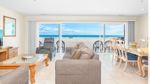 Ein geräumiges Wohnzimmer mit einer großen Schiebeglaswand, die auf einen Strand blickt, einem Essbereich mit Holzsesseln und einem Fernseher links.