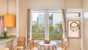 A dining area with a round table, two wooden chairs, a bowl of fruit, and a view of greenery through large windows.